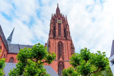 Frankfurt, Almanya - 10 Haziran 2025: Frankfurt Katedrali 'nin canlı yeşilliklerin ortasındaki yüksek Gotik kulesine yakından bakmak, bulutlu bir gökyüzüne karşı etkileyici mimariyi yakalamak,.