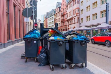 Frankfurt, Almanya - 10 Haziran 2025: Kentsel bir ortamda taşan çöp bidonları, tarihi yapılar ve açık mavi bir gökyüzüne karşı renkli atık sergiliyor.