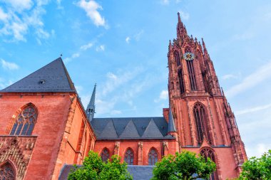 Frankfurt Katedrali 'nin yüksek kulesi ve çarpıcı kırmızı kumtaşı cephesinin detaylı görüntüsü, parlak gün ışığında ön planda yemyeşil ile güzelce aydınlatılmış..