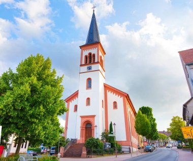 Rossdorf, Almanya - 23 Haziran 2025: Bir saat ve kule içeren uzun kilise kulesi, Rossdorf 'ta parçalı bulutlu bir gökyüzü altında yemyeşil ağaçlar ve yerleşim binaları ile çevrili.