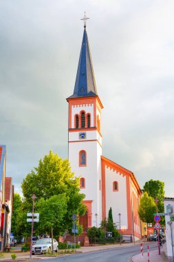 Rossdorf, Almanya - 23 Haziran 2025: Yeşillik ve konut binalarıyla çevrili tarihi bir kilise kulesinin büyüleyici manzarası Rossdorf 'ta karamsar, bulutlu bir gökyüzü altında.