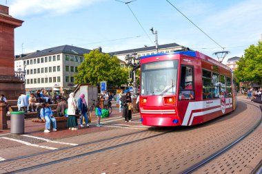 Darmstadt, Almanya - 5 Haziran 2025: Tramvay Almanya Darmstadt, Almanya 'da hareketli bir istasyona yaklaşıyor, bekleyen yolcular, canlı yeşil ağaçlar ve parlak mavi gökyüzü altında tarihi mimari.