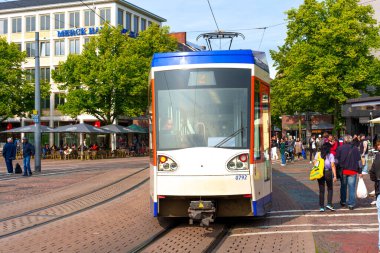 Darmstadt, Almanya - 5 Haziran 2025: Darmstadt 'ta yoğun bir istasyonda tramvay, açık mavi gökyüzünün altında bekleyen insanlar ve canlı yeşillikler.