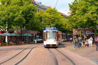 Darmstadt, Almanya - 5 Haziran 2025: Darmstadt, Almanya 'da yeşil ağaçlar ve güneşli bir günde kafelerin keyfini çıkaran yayalar tarafından çevrelenmiş canlı bir caddeye yaklaşan tramvay.
