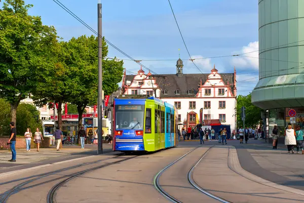 Darmstadt, Almanya - 5 Haziran 2025: Darmstadt, Almanya 'da tarihi mimari, ağaçlar ve yayalarla çevrili kalabalık bir meydanda raylar üzerinde hareket eden renkli tramvay.