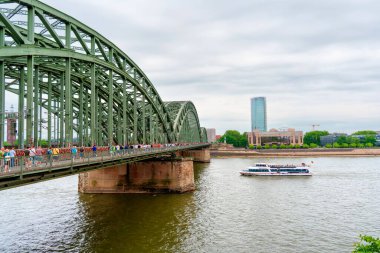 Köln, Almanya - 15 Haziran 2025: Hohenzollern Köprüsü, Köln 'deki Ren Nehri boyunca yayalar ve bulutlu gökyüzü altında bir nehir teknesinin yer aldığı, kentsel yaşamı ve mimariyi esir aldığı köprü..