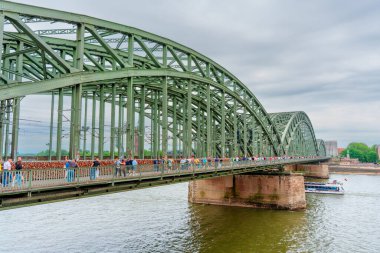 Köln, Almanya - 15 Haziran 2025: Köln 'deki Ren Nehri üzerindeki yaya köprüsü, bulutlu bir gökyüzü altında yemyeşil ve şehir ufuk çizgisini göstererek canlı aktiviteleri yakalıyor..