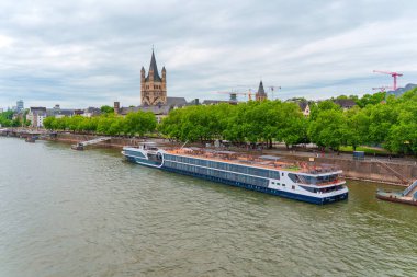 Köln, Almanya - 15 Haziran 2025: Ren nehrinin manzarasında büyük bir yolcu gemisi ve Köln silueti, bulutlu bir gökyüzüne karşı yeşil ağaçlar ve tarihi binalar sergileniyor..