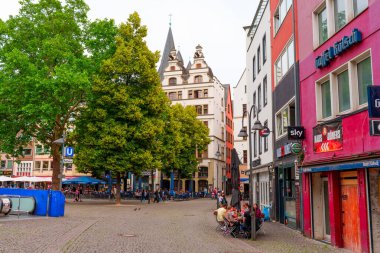 Köln, Almanya - 15 Haziran 2025: Cologne 'da tarihi binalar, açık kafeler, yemyeşil ağaçlar ve canlı atmosferin tadını çıkaran insanların yer aldığı canlı sokak manzarası.
