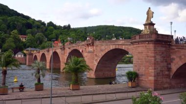 Heidelberg, Almanya - 21 Haziran 2025: İnsanlar Heidelberg 'in Eski Köprüsü' nde yürüyor ve bulutlu gökyüzünün altındaki palmiyeli nehir kıyısında oturuyor, tarihi köprüyü ve canlı sahneyi yakalıyorlar.
