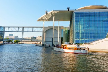 Berlin, Almanya - 16 Ağustos 2025: Berlin 'deki çağdaş cam binaların yakınında parlak mavi gökyüzünün altında süzülen beyaz tekne.