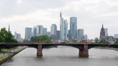 Frankfurt, Almanya - 10 Haziran 2025: Arka planda gökdelenleri olan Frankfurt Köprüsü, nehir kenarında yürüyen, koşan ve bisiklet kullanan insanlar, kentsel yaşam ve mimariye odaklı.