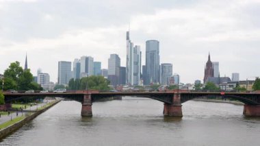 Frankfurt, Almanya - 10 Haziran 2025: Gökdelenli bir Frankfurt köprüsünün manzarası, nehir kenarında yürüyen, koşan ve bisikletle gezen insanları gösteriyor, kentsel yaşam ve mimariye odaklı.