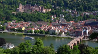 Heidelberg, Almanya - 21 Haziran 2025: Statik geniş çekim Heidelberg Kalesi 'ni yamaçta ve Eski Köprü' de turistlerle yürürken, Neckar Nehri 'nde bir açık turizm teknesi yelken açarken yakaladı..