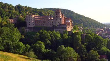 Heidelberg Şatosu bir tepenin üstünde duruyor, sıcak günbatımı ışığıyla yıkanmış, yemyeşil ağaçlarla çevrili, tarihi mimariyi gözler önüne seriyor..
