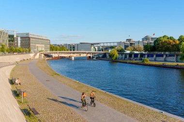 Berlin, Almanya - 16 Ağustos 2025: Spree Nehri 'nin yanındaki manzaralı bir yolda bisikletçiler, kent mimarisi içindeki eğlence faaliyetlerini vurguluyorlar.
