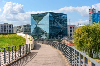 Berlin, Almanya - 17 Ağustos 2025: Küp Berlin cam yapısının modern tasarımı mavi gökyüzünü ve şehir manzarasını yansıtıyor, deniz boyunca bir yürüyüş yolu ile Berlin 'deki kentsel yaşamı yakalıyor.