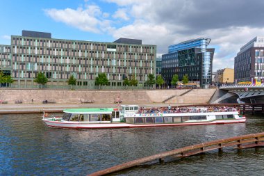 Berlin, Almanya - 17 Ağustos 2025: Spree Nehri üzerinde seyahat eden nehir teknesi, Berlin, Almanya 'da çağdaş binalar ve yemyeşil alanlarla çevrili.