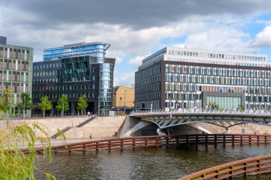 Berlin, Almanya - 17 Ağustos 2025: Berlin, Almanya 'da Veliaht Prens Köprüsü yakınlarındaki Spree Nehri boyunca uzanan çağdaş binalar, bulutlu bir gökyüzü altında cam ve taş tasarımlarının bir karışımı yer alıyor..