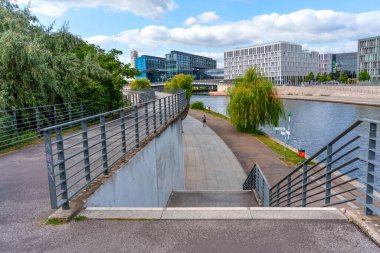 Berlin, Almanya - 17 Ağustos 2025: Güneşli bir günde arka planda modern mimari ile nehir kenarına giden yol.