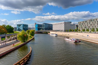 Berlin, Almanya - 17 Ağustos 2025: Berlin 'de berrak bir gökyüzü altında modern binalar, gezen tekneler ve yemyeşil yeşilliklerin yer aldığı Spree Nehri manzarası.