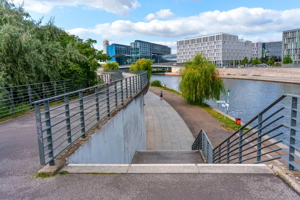 Berlin, Almanya - 17 Ağustos 2025: Güneşli bir günde arka planda modern mimari ile nehir kenarına giden yol.