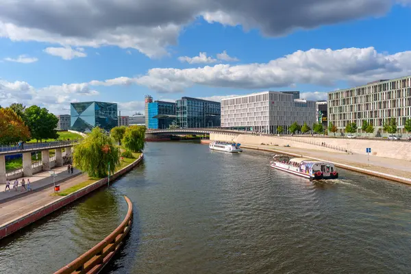 Berlin, Almanya - 17 Ağustos 2025: Berlin 'de berrak bir gökyüzü altında modern binalar, gezen tekneler ve yemyeşil yeşilliklerin yer aldığı Spree Nehri manzarası.