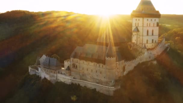 明るい太陽の光は 日没時に森林の丘の上に建てられた古代カルルシュタイン城を照らします チェコ共和国の歴史的建造物航空写真 — ストック動画