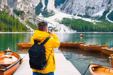 Braies gölünde tahta tekneler ve Dolomitlerin fotoğrafını çeken bir turist. Sarı ceketli ve siyah sırt çantalı bir adam İtalya 'da Dolomitleri ziyaret ediyor.. 