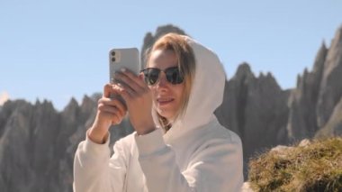 Güneş gözlüklü ve kapüşonlu gülümseyen kadın Tre Cime di Lavaredo yakınlarındaki dağların panoramik fotoğrafını çekiyor. Turist İtalyan Alplerini ziyaret etti