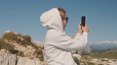 Sarışın kadın, Tre Cime di Lavaredo yakınlarındaki İtalyan Alplerindeki dağların telefonuyla fotoğraf çekiyor. Kapüşonlu turist gülümsüyor manzaraya hayran yavaş çekimde