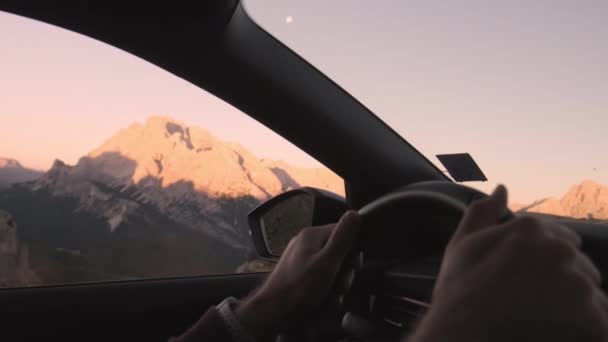 男は夜明けにイタリアアルプスの山の中で道路上の車を運転する 雲のない空の下で山脈を越えて運転する男性の手ターンステアリングホイールクローズアップ — ストック動画