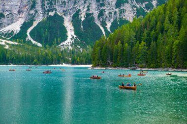 Turistler Braies gölünde ahşap kayıklara biniyorlar. Güneşli bir günde arkaplanda şeffaf su ve Dolomitler var..