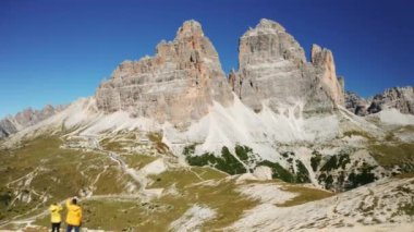 Birkaç genç turist Alplerdeki Tre Cime di Lavaredo 'ya bakıyor. Erkek ve kadın yüksek tepede, bulutsuz gökyüzü manzarasının altında dikiliyorlar.