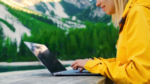 女性のフリーランサーは 森林の山や湖に対して木製のテーブルに座ってノートパソコン上のテキストを入力します 若い女性は新鮮な空気のクローズアップで作業を楽しんでいます — ストック動画