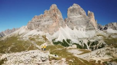 Kadın ve erkek uçurumun kenarındaki tepede durup görkemli dağlara bakıyorlar. Tre Cime di Lavaredo hava manzaralı geniş yeşil vadiler.