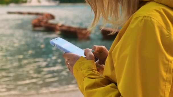 女性は背景のぼやけたスマートフォンでソーシャルネットワークをチェックします 女性ハイカーは 湖の近くに立ってガジェットを使用します — ストック動画