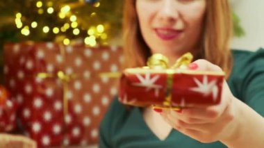 Yeşil elbiseli bir kadın ışıklarla, paketlenmiş hediyelerle ve arka planda bir Noel ağacıyla bir hediye verir.. 
