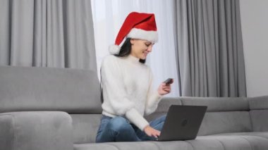 Kırmızı Noel Baba şapkalı bir kadın, oturma odasında oturan cep telefonu ve kredi kartıyla internetten Noel hediyesi alıyor.. 