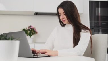 Moda mutfağında Not Defteri üzerine makale yazan odaklanmış genç bir kadın. Kadın serbest çalışan modern dizüstü bilgisayarda çalışıyor. Evde masada oturuyor.