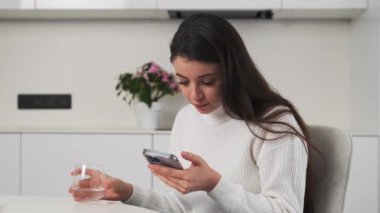 Odaklanmış kadın akıllı telefondan posta okurken su içer. Esmer kadın sabahları modern aletler kullanıyor şık mutfaktaki masada oturuyor.