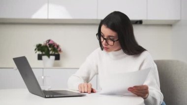 Gözlüklü esmer kadın deftere bakan kağıt belgelerdeki verileri kontrol ediyor. Odaklanmış genç çalışanlar şık mutfaktaki masada oturarak evde çalışırlar.