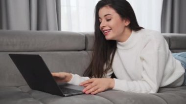 Neşeli kadın, oturma odasındaki kanepede uzanan Not Defteri aracılığıyla ailesiyle görüntülü konuşma yapıyor. Esmer kadın internet iletişimi için dizüstü bilgisayar kullanıyor.