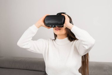 Woman puts on VR goggles to immerse into virtual reality. Young housewife sits on comfortable sofa using modern technology at home
