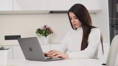 Satisfied woman types text on modern Notebook sitting at table in kitchen. Young freelancer enjoys working online using laptop slow motion