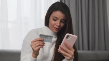 Young woman types details of credit card on smartphone to pay for purchase. Brunette lady makes online payment via gadget at home closeup