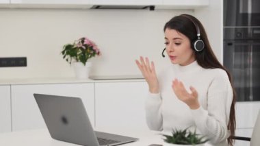 Female employee in headset talks to colleagues at online meeting via Notebook. Brunette woman explains project details at kitchen table slow motion