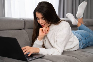 Happy young woman laying on sofa at home and looking on the laptop screen, reading news, watching video, chatting with friends