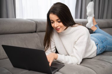 Young female laying on the sofa and using her notebook. A smiling woman has online shopping using a laptop at home. Working from home as a freelancer.