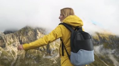 Happy woman raises hands contemplating view of Passo Giau pass covered with dense fog. Female tourist with backpack stands in Italian Alps slow motion